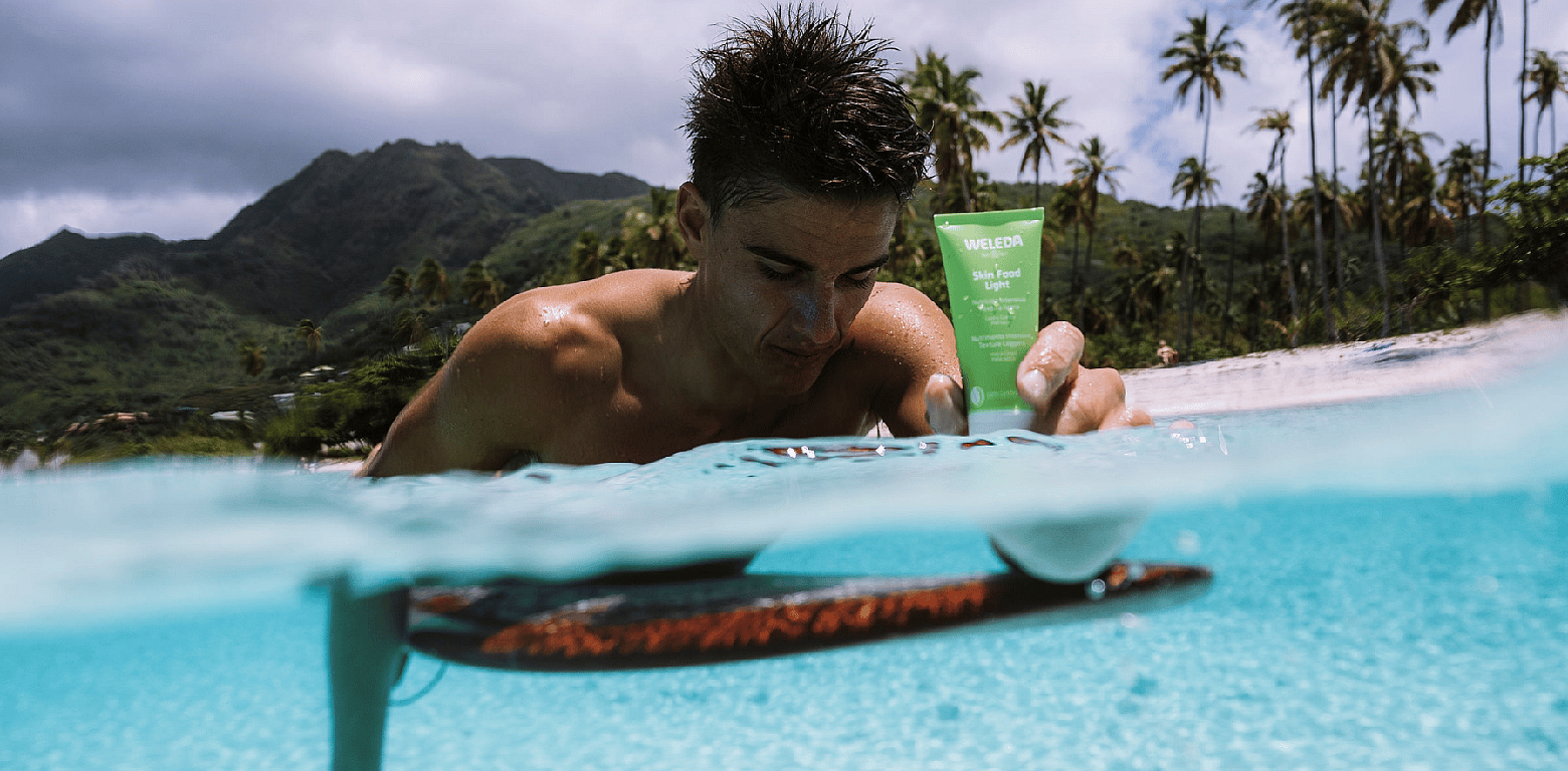 Hombre sobre una tabla de surf en el mar y sosteniendo una crema Skin Food de Weleda con la mano