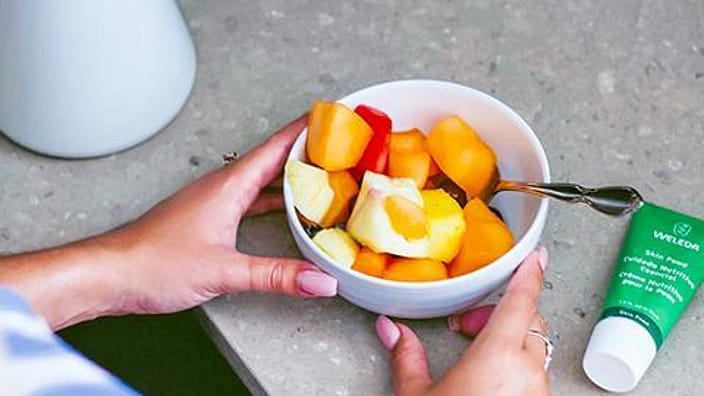 mujer sujetando un bol de frutas y de fondo la crema facial de Skin Food
